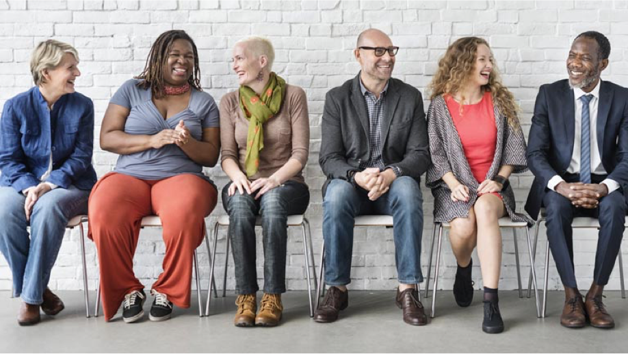 A group of 6 adults sitting in chairs in front of a white brick wall talking.