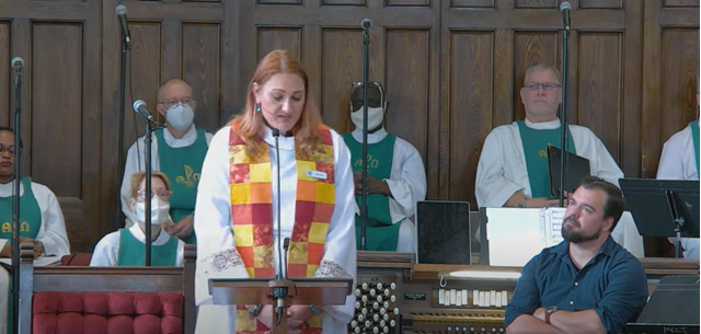 Picture of Rev. Junia Joplin preaching at the Sanctuary, with the Choir of MCC Toronto sitting behind her.