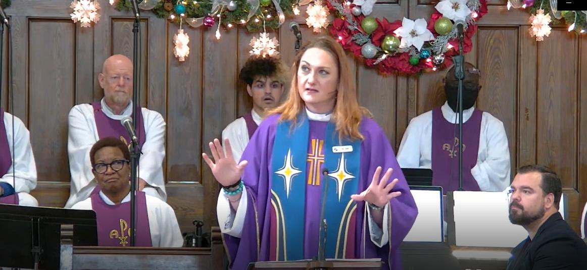 Picture of Rev. Junia Joline preaching, with the Choir of MCC Toronto members behind her.