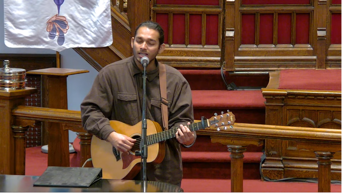 Picture of our offertory performer Just Prince, holding a guitar and singing into a mic.