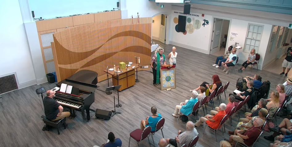 Ariel shot of MCC Toronto's Sunday Service in the social hall, with Rev. Deana and Sheryl Pollack at the podium, Music Director Jason Jesdadt at the Piano in front of the seated congregants.