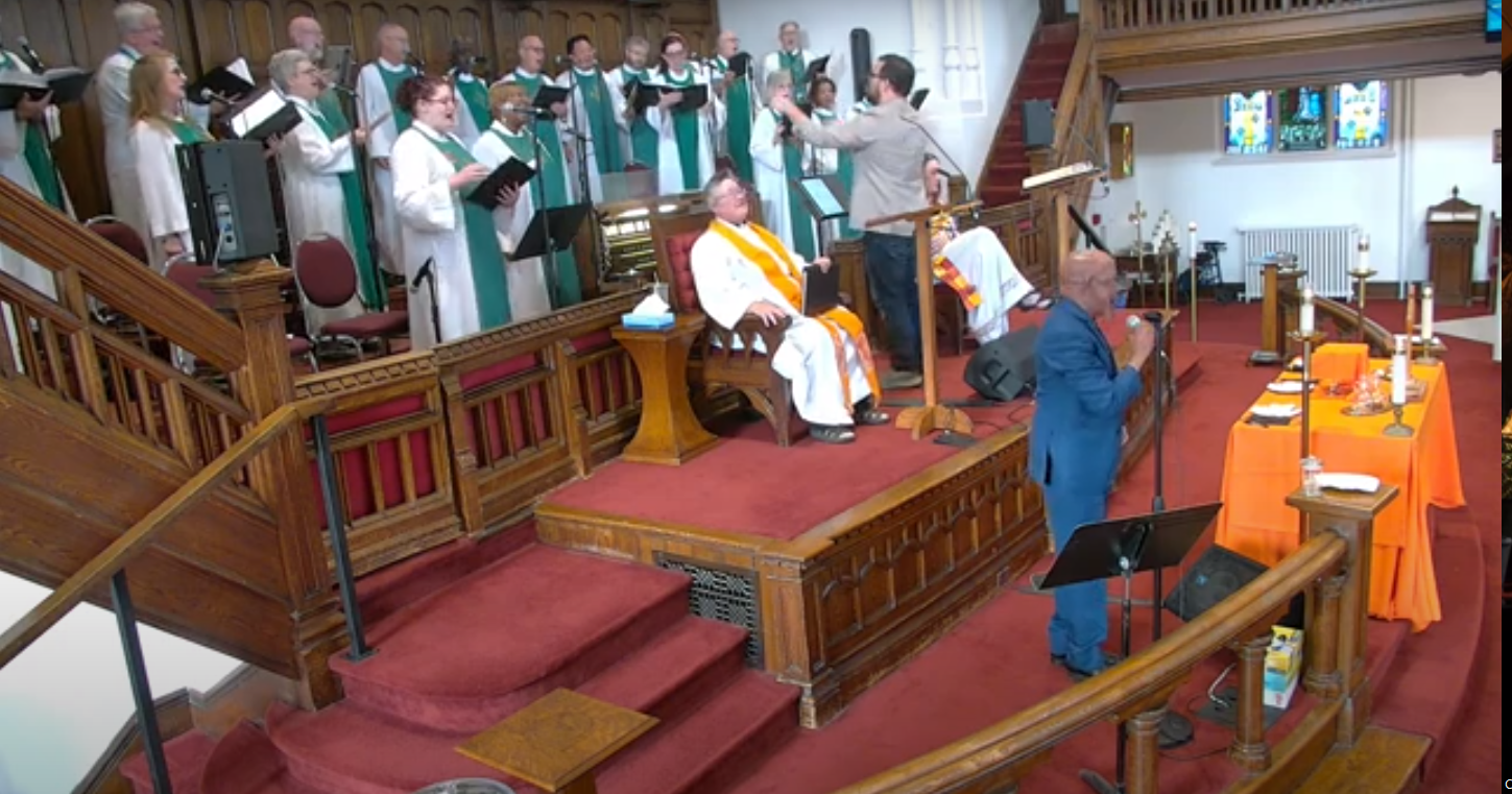Image of the Sunday Service at the Sanctuary with The Choir of MCC Toronto singing, Rev. Deana seated in front of them, and Marl Cassius singing in front of them all.